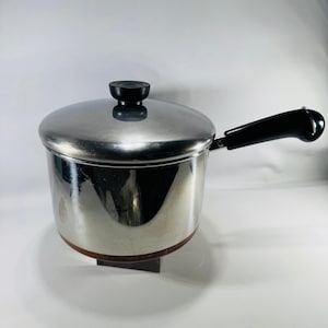 May include: A stainless steel saucepan with a polished finish, featuring a matching lid with a black knob and a long, curved black handle. The base of the pot has a copper-colored bottom, and the entire piece has a reflective, metallic surface.