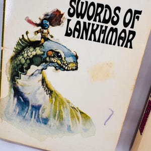 May include: Vintage paperback book cover featuring the title "Swords of Lankhmar" in black lettering. The cover art depicts a fantastical creature with a rider, rendered in shades of blue, green, and brown. The overall aesthetic is reminiscent of classic fantasy illustrations.