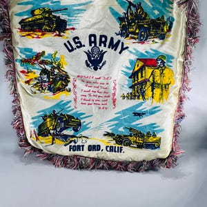 May include: Cream-colored pillow with a U.S. Army theme, featuring illustrations of military vehicles and soldiers. The pillow has a decorative fringe and includes text that reads "U.S. ARMY" and "FORT ORD, CALIF."