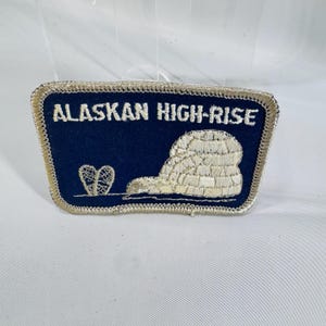 May include: Embroidered patch with the text "ALASKAN HIGH-RISE" in white on a navy blue background. The patch features a silver-colored igloo and a heart-shaped design. The patch has a silver border.