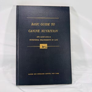 May include: A hardcover book titled "BASIC GUIDE TO CANINE NUTRITION" with a special section on "NUTRITIONAL REQUIREMENTS OF CATS." The cover is dark blue with gold lettering and a small illustration of a dog. The book is from the Gaines Dog Research Center, New York.