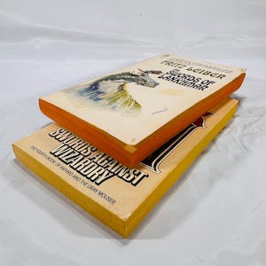 May include: Two vintage paperback books. The top book is titled "Swords of Lankhmar" by Fritz Leiber, featuring a dragon illustration. The bottom book is "Swords Against Wizardry". Both books have orange and yellow edges.