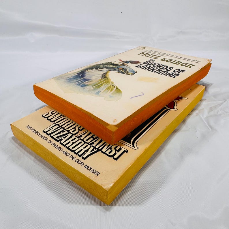 May include: Two vintage paperback books. The top book is titled "Swords of Lankhmar" by Fritz Leiber, featuring a dragon illustration. The bottom book is "Swords Against Wizardry". Both books have orange and yellow edges.