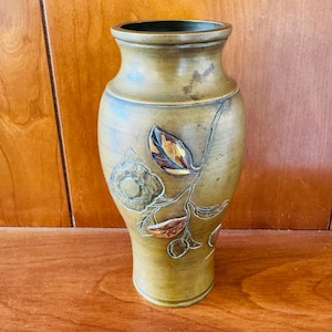 May include: A decorative vase with a floral design. The vase is a muted green color with a dark green rim. The floral design features a flower and leaves in shades of gold, copper, and brown.