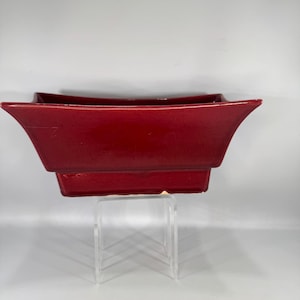 May include: A glossy, deep red ceramic planter with a rectangular shape and flared sides. The planter has a tiered base and is displayed on a clear acrylic stand. The design is modern and minimalist.