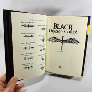 May include: An open book titled "Black Dragon Codex" by R.D. Henham. The book's pages are cream-coloured with black text and illustrations of dragons. The left page lists other dragon codices. The book is bound in a dark brown cover.