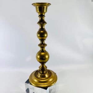 May include: A tall, polished brass candlestick holder featuring a wide, circular base, a slender central stem with two prominent spherical nodes, and a flared top cup designed to securely hold a single taper candle for display purposes.