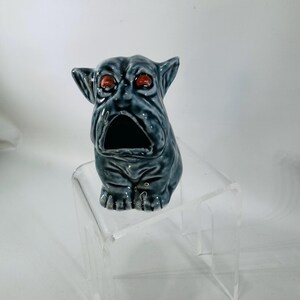 May include: A ceramic figurine of a bulldog-like creature with a wide open mouth and red eyes. The figurine is a dark blue-grey color and sits on a clear acrylic stand. The figurine has a glossy finish.