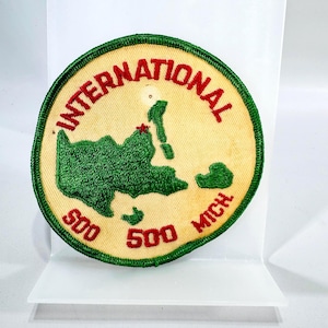 May include: A round patch with a cream-colored background and green and red embroidered details. The patch features the word "INTERNATIONAL" in red, a green map, and the words "500 MICH" and "500" in red.