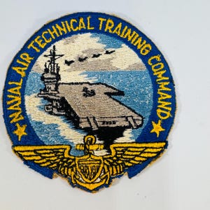 May include: Embroidered patch featuring a navy aircraft carrier on a blue and white background. The patch has the text "NAVAL AIR TECHNICAL TRAINING COMMAND" in a circular design. A gold emblem with wings and an anchor is at the bottom.