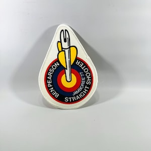 May include: A white, tear-drop shaped patch with a target design and the text "BEN PEARSON SHOOTER STRAIGHT JAMBOREE 77". The target has red, yellow, and blue concentric circles. A yellow arrow is in the center.