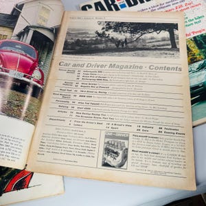 May include: A vintage "Car and Driver Magazine" open to its contents page, with a red car visible on the left. The magazine features articles on racing and car reviews. The cover has a black and white landscape photo.