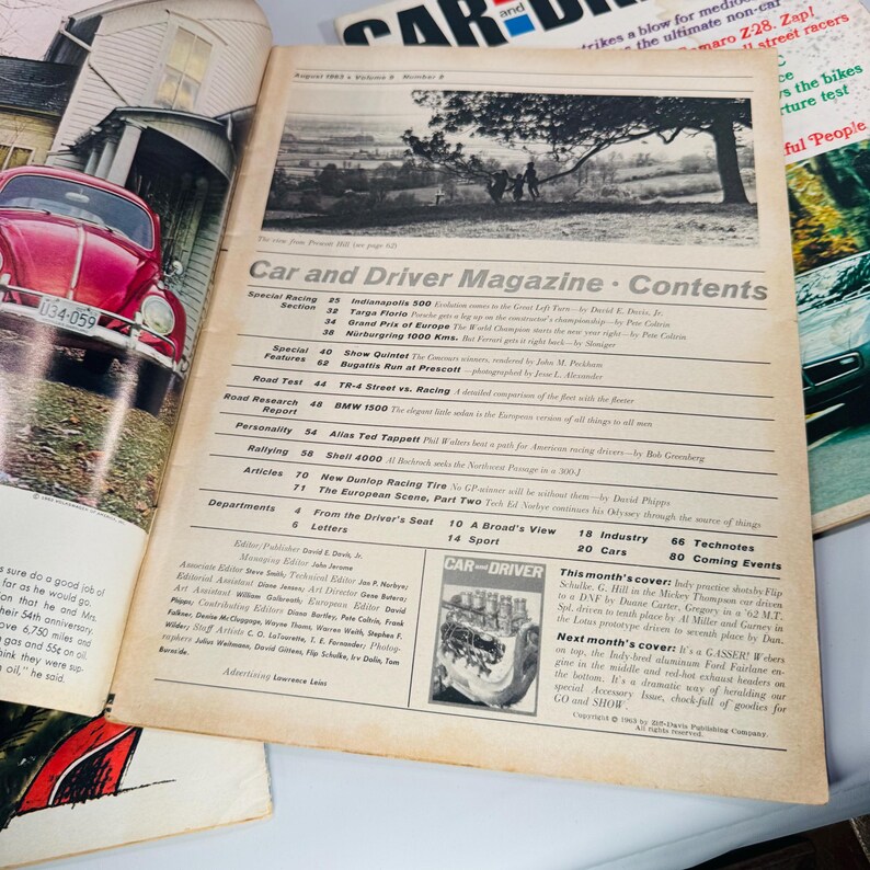 May include: A vintage "Car and Driver Magazine" open to its contents page, with a red car visible on the left. The magazine features articles on racing and car reviews. The cover has a black and white landscape photo.
