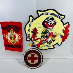 May include: A collection of embroidered patches. One red patch reads "Emergency Service Explorer." A yellow patch features a cartoon firefighter with the text "Fire Department Staff." A circular patch displays the American Red Cross logo.