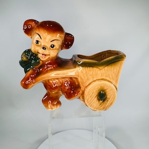 May include: A vintage ceramic planter shaped like a brown bear pulling a tan wagon. The bear has a smiling face, dark eyes, and a green bow. The wagon has a green trim and a single wheel.