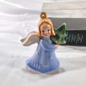 May include: A vintage ceramic angel ornament from 1979. The angel has blonde hair, a blue dress with star accents, and white wings. She holds a green Christmas tree. The ornament is approximately 3 inches tall.