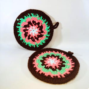May include: Two round, handmade crochet potholders. The potholders feature a brown outer ring, with concentric circles of green, pink, and white. One potholder has a small loop for hanging. The potholders are on a white background.