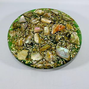 May include: A round, decorative coaster with a green border. The coaster is filled with small, tan pebbles and iridescent shell fragments. The shells have shades of pink, orange, and gold. The coaster is a unique home decor item.