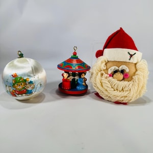 May include: Three Christmas ornaments: a white satin ball with a cat illustration, a miniature carousel with figures, and a Santa Claus head with sunglasses. The ornaments are festive and colorful, perfect for holiday decorating.