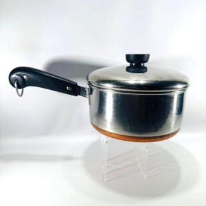 May include: A stainless steel saucepan with a lid and a black handle. The pot has a wooden base and a black knob on the lid. The handle has a metal loop. The pot is on a clear stand.