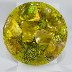 May include: A round, decorative resin piece with a vibrant yellow-green hue. Embedded within are iridescent abalone shells and sparkling glitter. The shells vary in size and shape, creating a unique, eye-catching design.