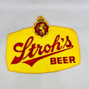 May include: A yellow embroidered patch featuring the red text Stroh's BEER in a script font with a red and yellow crest above it. The crest displays a lion figure and a crown. The patch has a yellow border and a textured fabric surface.