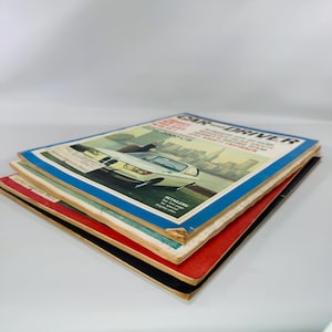 May include: A stack of vintage Car and Driver magazines. The top magazine has a blue border and features a white car with a cityscape background. The other magazines have red and black covers. The magazines are stacked on a white surface.
