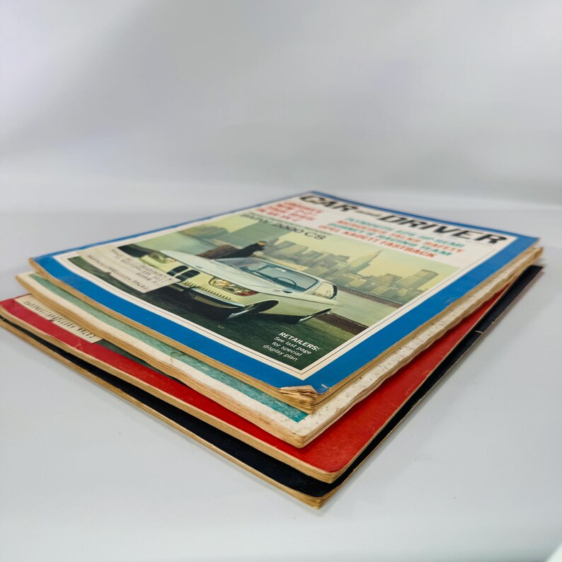 May include: A stack of vintage Car and Driver magazines. The top magazine has a blue border and features a white car with a cityscape background. The other magazines have red and black covers. The magazines are stacked on a white surface.