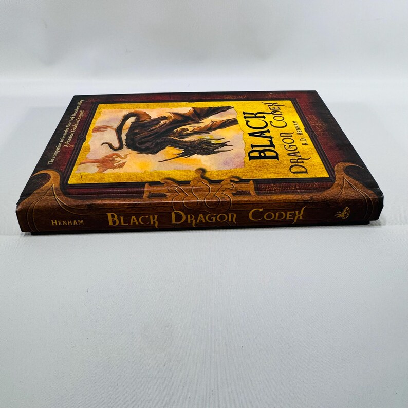 May include: A hardback book titled "Black Dragon Codex" by R.D. Henham. The cover displays a black dragon illustration on a yellow background, framed by a dark brown border. The book's title is prominently displayed in gold lettering.
