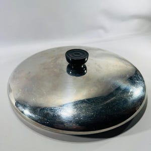 May include: A circular, polished stainless steel pot lid featuring a smooth, reflective surface and a centered, black, circular knob handle with concentric ring detailing on its top. The lid has a slightly domed shape and a clean, metallic finish.