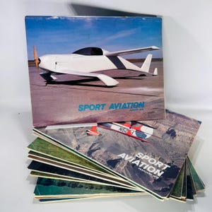 May include: A stack of vintage "Sport Aviation" magazines. The top magazine features a white airplane with a propeller against a blue sky. The text "SPORT AVIATION" and "JANUARY 1976" are visible. Other magazines show aerial views.