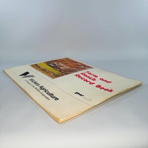 Pu&ograve; includere: Un vecchio "Farm and Ranch Record Book" con un'immagine di paesaggio sulla copertina. La copertina presenta anche il testo "Wickes Agriculture" e "A Division of the Wickes Corporation". Il titolo "Farm and Ranch Record Book" &egrave; in rosso.