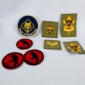 May include: A collection of vintage Boy Scout patches. The patches feature various designs, including a tiger, a raven, and the Boy Scouts of America emblem. Colors include navy blue, red, yellow, and green. The patches are made of fabric and have embroidered details.