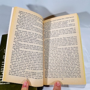 May include: An open book with yellowed pages, featuring text and the numbers 32 and 33. The book is held open by a person's fingers, with a dark green book visible in the background.