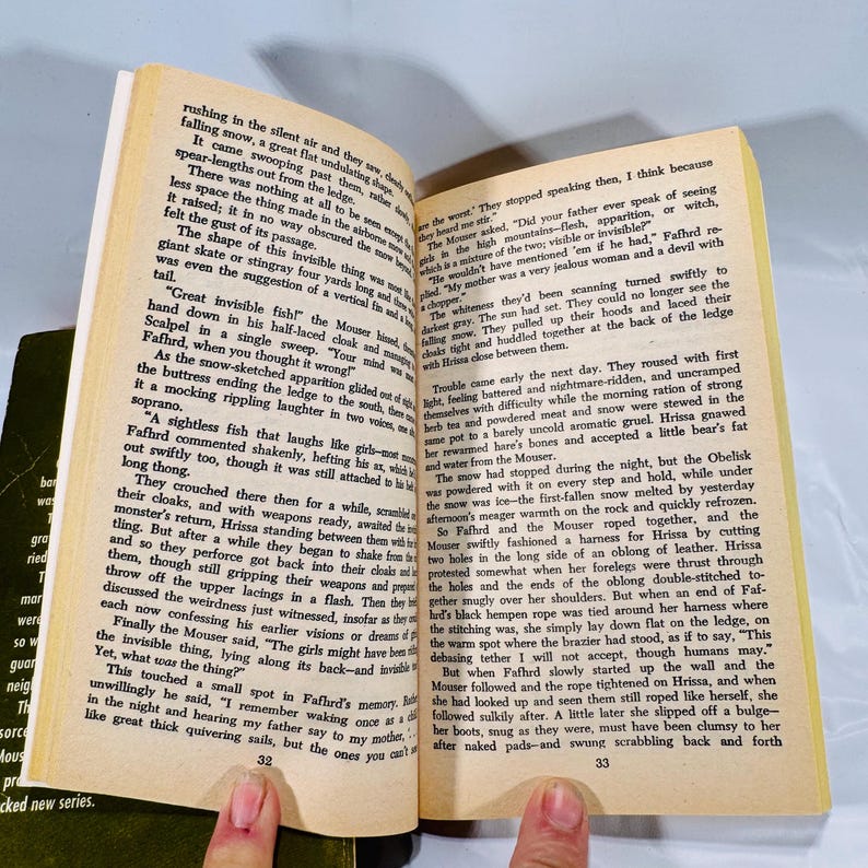 May include: An open book with yellowed pages, featuring text and the numbers 32 and 33. The book is held open by a person's fingers, with a dark green book visible in the background.