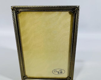 May include: A rectangular brass-toned picture frame with a decorative rope-like border. The frame holds a yellowed paper. A small oval sticker with text and the price $5.59 is on the bottom right. The frame is 3.5 x 5 inches.