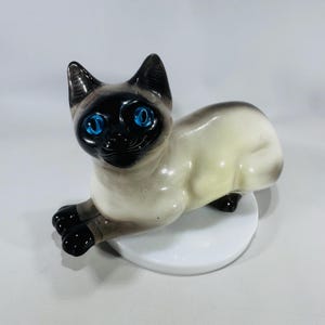 May include: A ceramic Siamese cat figurine with striking blue eyes. The cat is in a reclining pose, with black ears, paws, and face, and a cream-colored body. It rests on a white, circular base.