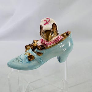 May include: A light blue ceramic shoe figurine with a brown mouse wearing a pink and white plaid scarf and a white hat with a red number 2. The shoe has a high heel and decorative details.