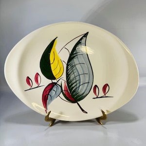 May include: An oval-shaped cream-colored ceramic platter featuring a hand-painted botanical design. The pattern includes large stylized leaves in shades of yellow, green, and blue, accented by smaller red leaf motifs and thin black lines.