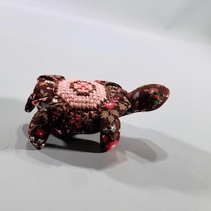May include: A handmade fabric turtle toy. The turtle's body is brown with a floral pattern in red, pink, and white. The turtle's shell is a light pink with a crocheted design. The head has a red and green detail.