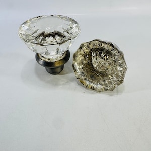 May include: Two clear glass knobs with a faceted, multi-sided design. One knob is attached to a dark metallic base, while the other is shown separately, displaying its textured, star-like pattern on the underside of the glass surface.