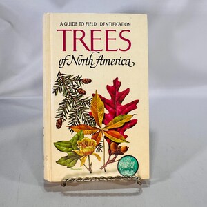 Puede incluir: Un libro antiguo titulado "TREES of North America" con una cubierta color crema. La cubierta presenta ilustraciones de varias hojas, piñas y una flor. El libro forma parte de la serie "Golden Field Guide".