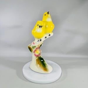 May include: A ceramic figurine of two yellow birds perched on a branch with a pink flower. The birds have black eyes and are facing different directions. The branch is white with black dots. The base is white and the figurine is on a white surface.
