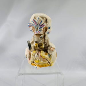 May include: A ceramic figurine of a mouse wearing a bonnet and holding a small mirror. The mouse is dressed in a cream-colored outfit with yellow and blue accents. The bonnet has pink and blue decorative elements.