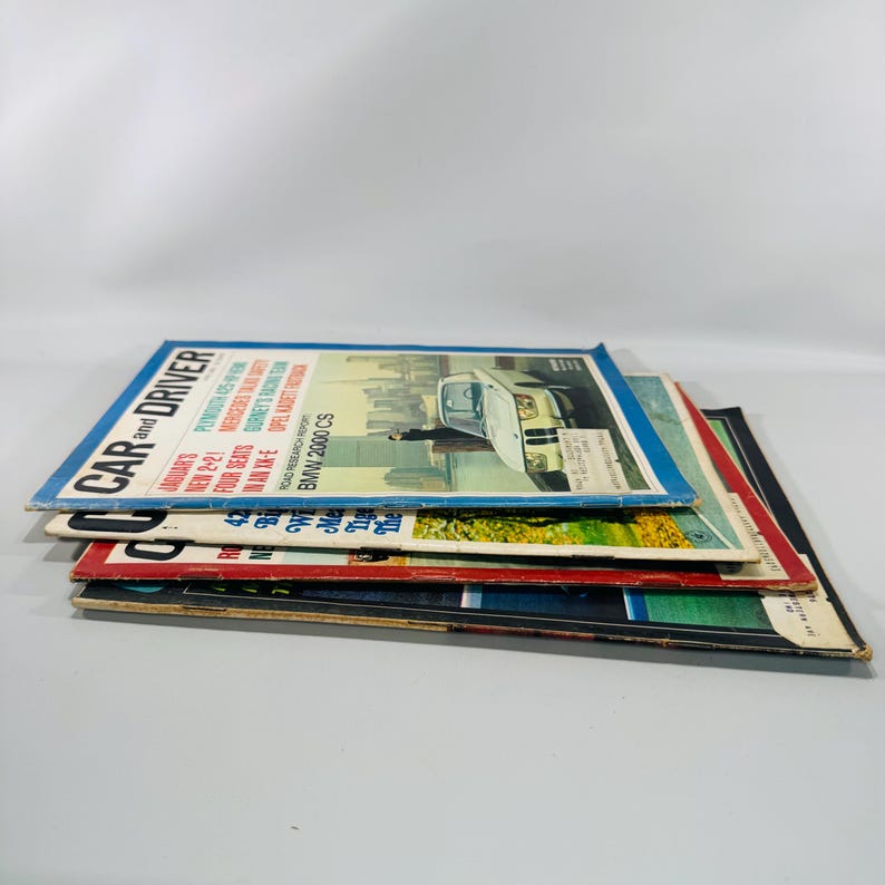 May include: A pile of vintage "Car and Driver" magazines. The top magazine shows a car on the cover with the text "BMW 2000 CS". The magazines have a blue, red, and black colour scheme.