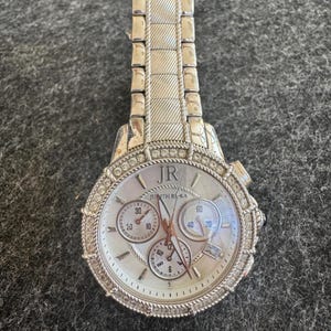 May include: Silver-toned wristwatch with a textured band and a round face. The watch face features three sub-dials and is encrusted with small, clear crystals around the bezel. The brand name "Judith Rl Ka" is visible on the watch face.
