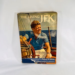 May include: A vintage hardcover book titled "The Living JFK" with a photo of John F. Kennedy on a sailboat. The book cover features a blue and white color scheme with a red title and a blue background.