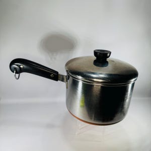 May include: Stainless steel saucepan with a black handle and lid. The pot has a copper-colored bottom and a small loop on the handle. The lid has a black knob. The saucepan is designed for cooking.