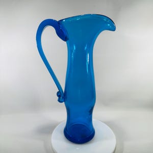 May include: A tall, vibrant blue glass pitcher with a curved handle. The pitcher has a flared top and a rounded base. The handle is attached near the top and bottom of the pitcher. The pitcher is displayed on a white surface.