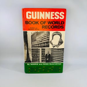 May include: A vintage Guinness Book of World Records. The cover is orange and white with the title in large white letters. The bottom of the book is green with a list of superlatives. The book features black and white images.